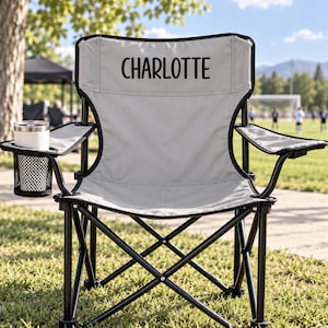 May include: A light grey folding camping chair with a black metal frame and a mesh cup holder on the left armrest. The backrest features the name CHARLOTTE printed in bold black capital letters. The chair has a collapsible design.