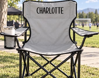 May include: A light grey folding camping chair with a black metal frame and a mesh cup holder on the left armrest. The backrest features the name CHARLOTTE printed in bold black capital letters. The chair has a collapsible design.