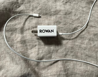 Op de afbeelding: Een witte wandadapter met het woord ROWAN in zwarte letters op de zijkant. De adapter is voorzien van een tweepolige stekker en is verbonden met een lange witte oplaadkabel met een lightning-connector aan het uiteinde van het snoer.