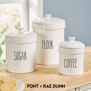 May include: Three white ceramic canisters with black lids and labels. The labels read "Sugar", "Flour", and "Coffee". The canisters are on a wooden countertop. The font is Rae Dunn.