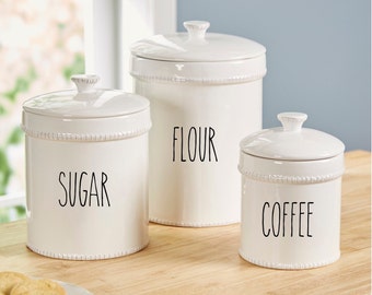 May include: Three white ceramic canisters with lids, each featuring a textured beaded rim and black lettering. The canisters are labeled SUGAR, FLOUR, and COFFEE in a tall, thin font. The containers have a glossy finish and matching lids.