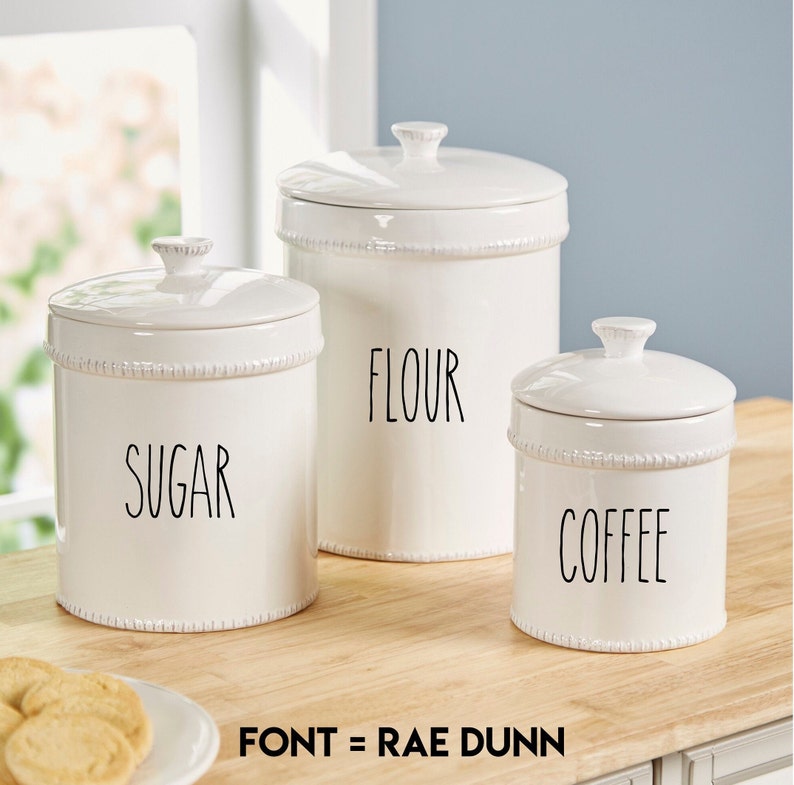 May include: Three white ceramic canisters with black lids and labels. The labels read "Sugar", "Flour", and "Coffee". The canisters are on a wooden countertop. The font is Rae Dunn.