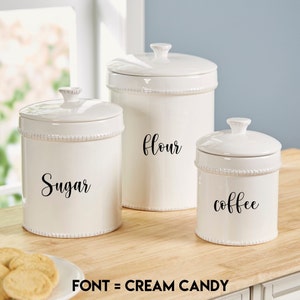 May include: Three white ceramic canisters with black lettering. The canisters are labeled "Sugar", "Flour", and "Coffee".