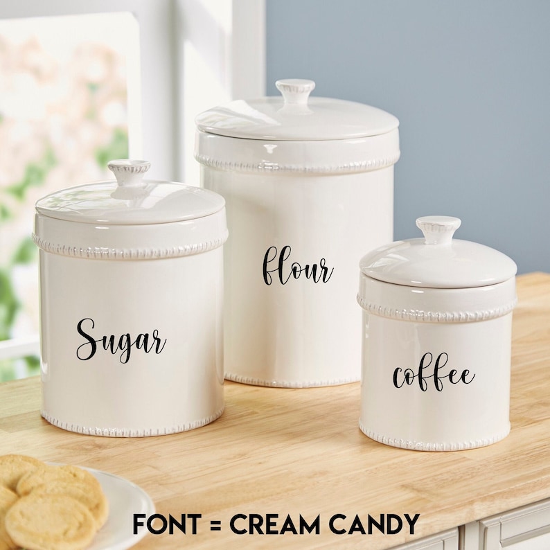 May include: Three white ceramic canisters with black lettering. The canisters are labeled "Sugar", "Flour", and "Coffee".