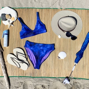 A styled flat lay of both the top and bottom together outside in natural light at the beach. The Bikini top and bottom are a vibrant purple with blue touches. The lining is purple. Top and bottom are sold separately.