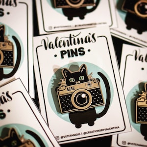 May include: Enamel pin featuring a black cat with green eyes, integrated into a camera design. The pin is on a white card with the text "Valentina's PINS" and a light blue circle background. The pin is black and gold.