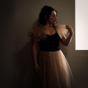 Puede incluir: Un top tipo corsé negro combinado con una falda de tul beige con lentejuelas dispersas. El conjunto incluye un cuello de tul beige a juego con adornos de lentejuelas, creando un aspecto en capas con un corpiño oscuro y una falda brillante.