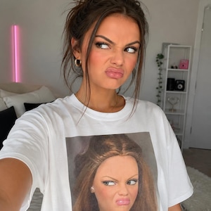 May include: A white short-sleeved t-shirt featuring a square graphic print on the chest. The print displays a close-up portrait of a doll with long brown hair, arched eyebrows, and a skeptical facial expression, mirroring the wearer's expression.
