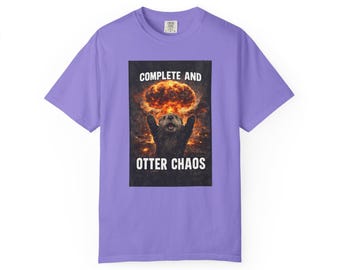Puede incluir: Una camiseta de color lila con un gráfico de una nutria con los brazos levantados frente a una nube en forma de hongo. El texto "COMPLETE AND OTTER CHAOS" está impreso encima y debajo de la imagen de la nutria en blanco.