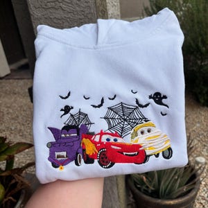 K&ouml;nnte beinhalten: Wei&szlig;er Kapuzenpullover mit einem gestickten Halloween-Design. Das Design zeigt drei Zeichentrickautos, die als Vampir, Mumie und Rennwagen verkleidet sind, mit Flederm&auml;usen, Spinnennetzen und Geistern dar&uuml;ber.