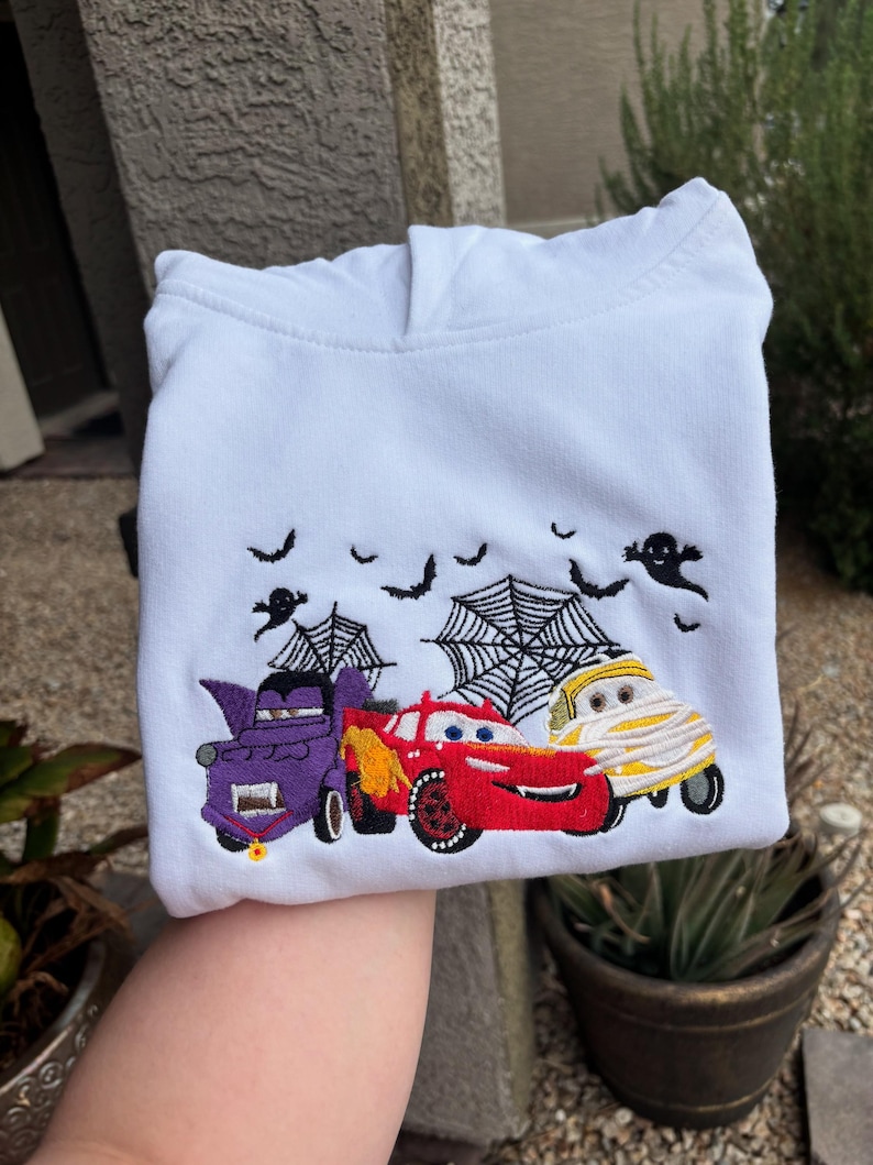 K&ouml;nnte beinhalten: Wei&szlig;er Kapuzenpullover mit einem gestickten Halloween-Design. Das Design zeigt drei Zeichentrickautos, die als Vampir, Mumie und Rennwagen verkleidet sind, mit Flederm&auml;usen, Spinnennetzen und Geistern dar&uuml;ber.