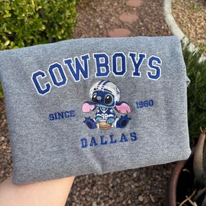 Puede incluir: Sudadera gris con la palabra "COWBOYS" en letras azules. Debajo, un bordado de un personaje de dibujos animados con casco de fútbol y balón. Las palabras "SINCE 1960" y "DALLAS" también están bordadas.