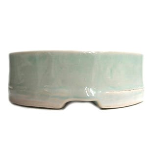 May include: A pale green ceramic bonsai pot with a rounded rectangular form. The pot has a smooth, glazed surface and a white base with two cut-out sections. The pot is designed for holding a small plant.