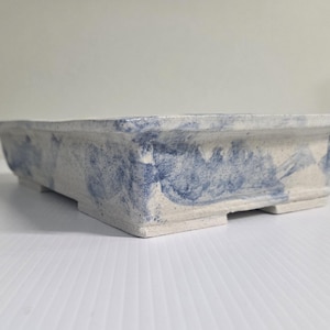 Peut inclure: Un pot à bonsaï rectangulaire en céramique avec une base texturée blanc cassé et des motifs irréguliers de coups de pinceau bleus sur les côtés extérieurs. Le récipient présente un large rebord, des parois légèrement inclinées et quatre petits pieds.