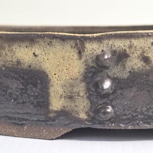 May include: A shallow ceramic tray with a dark brown, textured base and a contrasting cream-coloured glaze dripping down the sides. The rim features three small, raised, rounded bumps arranged vertically along the exterior surface.
