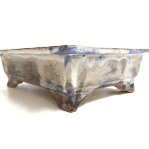 May include: A square ceramic bonsai pot with a light blue rim and a textured, mottled surface in shades of grey, white, and brown. The pot has small, triangular feet, designed for bonsai trees.