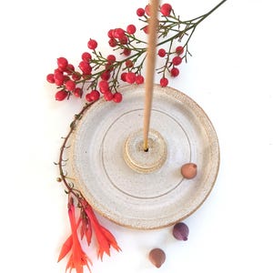 May include: A light beige ceramic incense holder with a central hole for the incense stick and concentric circles. Red berries and orange flowers are arranged around the holder, with a few small decorative stones. The holder is handmade.