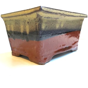 May include: A square bonsai pot made of ceramic, featuring a layered colour scheme. The top section is yellow and brown, transitioning to black, and a deep red base. The pot has small feet and a slightly flared shape.