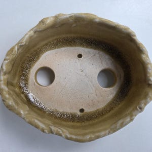 May include: An oval ceramic bonsai pot with a textured, uneven rim. The interior features a light beige base colour with a dark brown, speckled glaze gradient along the inner walls. Two large circular drainage holes and two smaller holes are visible.