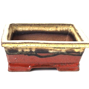 May include: A square ceramic bonsai pot with a red, brown and yellow glaze. The pot has a rectangular shape with four small feet.
