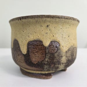 May include: A small ceramic cup with a rounded shape and a wide rim. The exterior features a textured, speckled beige glaze on the upper portion that drips down over a dark, mottled brown base. The surface has a porous, earthy appearance.