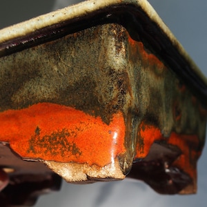 May include: A square ceramic bonsai pot with a dark green, brown, and orange glaze. The pot has a rough, textured surface and a slightly irregular shape.