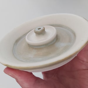 May include: A ceramic incense holder with a white and pale green glaze. The holder has a circular base with a raised central section for incense sticks. The design is simple and elegant, suitable for home decor.
