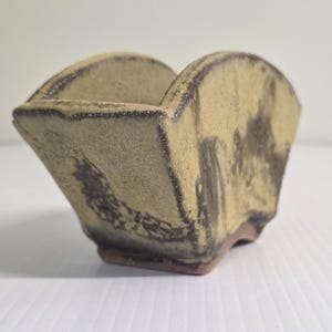 May include: A hand-thrown ceramic coffee dripper with a textured, speckled beige finish and dark brown glazed edges. The vessel features a wide, flared top opening and a tapered base with a distinct, unglazed reddish-brown clay foot.