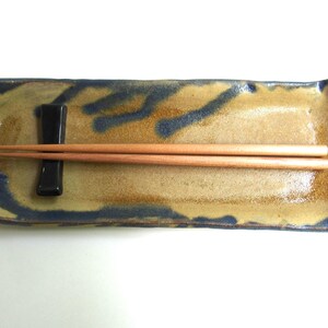 May include: A rectangular ceramic dish with a blue and brown glaze. The dish has a small black ceramic rest for chopsticks. A pair of wooden chopsticks are resting on the dish.