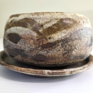 May include: A small, round ceramic bowl with a matching saucer, featuring a mottled, earthy glaze in shades of cream, beige, and dark brown. The surface has a textured, uneven appearance with swirling, organic patterns across the exterior.