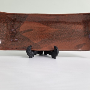 May include: A rectangular ceramic tray with a mottled reddish-brown glaze and a glossy finish. The tray features slightly curved edges and is displayed on a black plastic stand. The surface has a textured appearance with darker brown speckles.