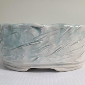 May include: A round ceramic bowl with a textured, wavy surface featuring a glossy white glaze with subtle light blue streaks. The bowl has an uneven, organic rim and a slightly raised base, showcasing a hand-formed appearance with visible ridges.