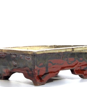 Könnte beinhalten: Ein rechteckiger Bonsai-Topf mit dunkelbrauner Glasur und einem hellbraunen Rand. Der Topf hat vier Beine und eine rot-braune Glasur auf der Unterseite.