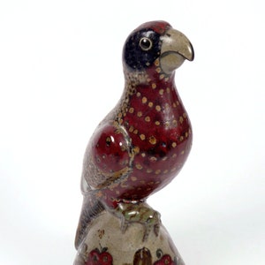 Può includere: Una statuetta di uccello in ceramica con vernice macchiettata di rosso, marrone e bianco. L'uccello è appollaiato su una base con un disegno floreale.