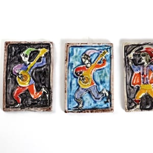 May include: Three ceramic wall plaques depicting figures playing musical instruments. The figures are dressed in colorful clothing and have exaggerated features. The plaques are rectangular and have a black background.