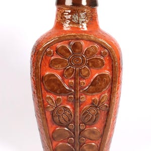 Op de afbeelding: Een oranje keramische vaas met een bruin bloemmotief. De vaas heeft een getextureerd oppervlak en een smalle hals. Het bloemmotief is symmetrisch, met een grote bloem bovenaan en kleinere bloemen en bladeren eronder. Ongeveer 30 cm hoog.