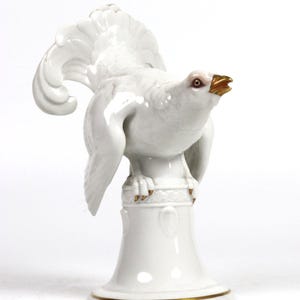 Op de afbeelding: Een wit porseleinen beeldje van een vogel met een gouden snavel en accenten. De vogel zit op een wit voetstuk met gouden versieringen.