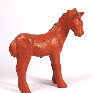 Puede incluir: Una pequeña figura de caballo de color rojo anaranjado sobre un fondo blanco. El caballo de juguete tiene una superficie texturizada con detalles verdes en la crin y el cuerpo. El caballo es de color sólido y tiene un diseño de juguete clásico.