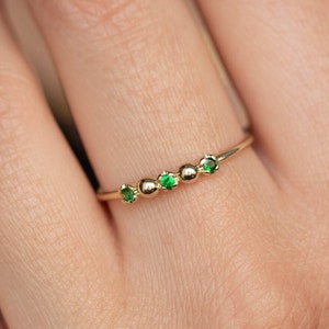 Puede incluir: Un delicado anillo de oro con cinco pequeñas piedras preciosas verdes dispuestas en fila. El anillo se lleva en un dedo.