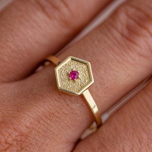 May include: A gold ring with a hexagonal setting featuring a small red gemstone. The ring has a textured surface.