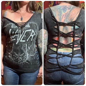 May include: A black Slayer band t-shirt with a distressed design and the band's logo in white. The shirt has a lace-up neckline and cut-out detailing on the sides. The back of the shirt features a woven design. The model is wearing blue jeans.