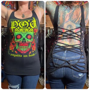 May include: Black tank top with a graphic of a skull with red eyes, green and yellow accents, and the text "payable on death." The back of the tank top has a crisscross design. The person is wearing blue jeans.