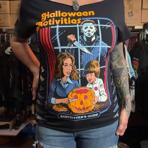 May include: Black off-the-shoulder t-shirt featuring a Halloween-themed graphic. The graphic includes the text "Halloween Activities" and "Babysitter's Guide", along with illustrations of a jack-o'-lantern and a figure holding a knife.