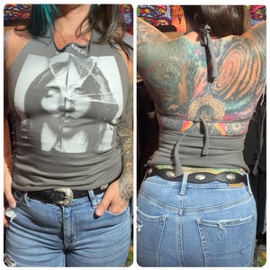 May include: A gray sleeveless halter top featuring a black and white graphic print of a face on the front. The top has a tie-neck closure and a tie-back design, paired with light blue denim jeans and a black belt with a silver-toned buckle.