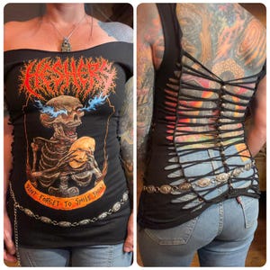 May include: A black off-the-shoulder top featuring a graphic of a skeleton holding a mask with blue flames from its eyes. The text reads HESHERS and DONT FORGET TO SMILE TODAY. The back has a lattice design with silver beads and a silver chain belt.