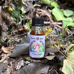 May include: A small, clear glass bottle with a black cap, containing a brown liquid. The bottle is labeled "Sun's Eye" and "Vision Quest Concentrated Aroma 1/2 fl oz (15 ml)". The bottle is sitting on a bed of green leaves.