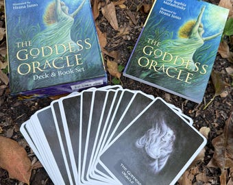 May include: A deck of oracle cards with a black and white design. The cards are fanned out on a bed of brown leaves. The box for the cards is visible in the background. The box is blue and green with the text "The Goddess Oracle Deck & Book Set" printed on the front.