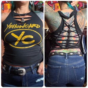May include: A black sleeveless tank top featuring the yellow text "YELLOWCARD" and a "YC" logo on the front. The garment has horizontal cutouts at the neckline and a complex, woven, lattice-style cutout design across the entire back panel.