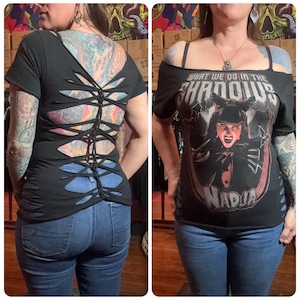 May include: Black t-shirt with cut-out back and shoulder details. The front features a graphic of a woman with the text "WHAT WE DO IN THE SHADOWS NADJA". The shirt is paired with blue jeans.