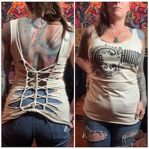 May include: Beige tank top with a unique cut-out design on the back, laced up with a criss-cross pattern. The front features the word "HIDDEN" in a decorative black font. The tank top is paired with blue jeans.